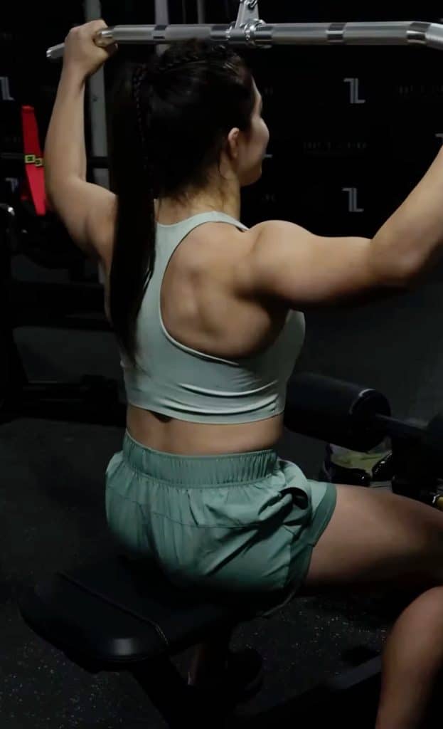 A woman wearing GLYDE Shorts uses a lat pulldown machine at the gym, viewed from behind, showcasing her strong back and arms.