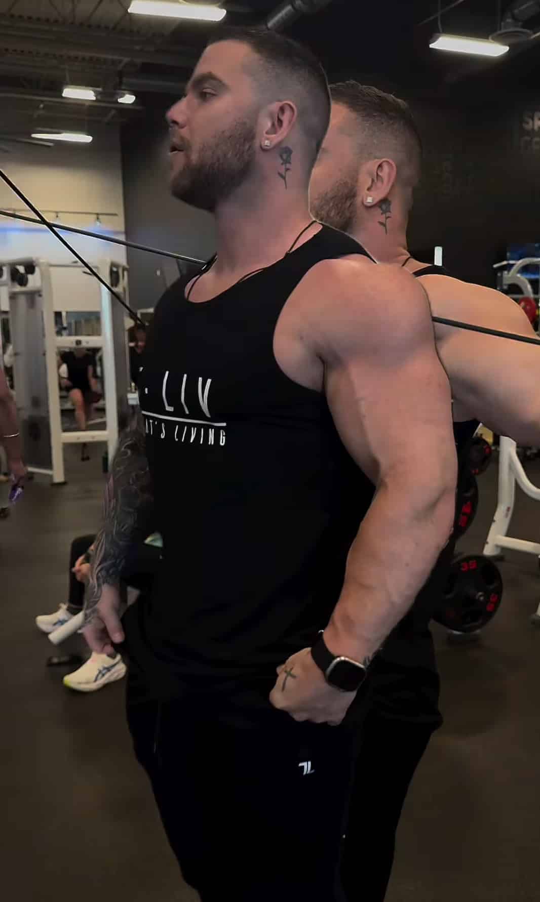 Two muscular men stand back-to-back in a gym, both wearing the Genesis Tank. One assists the other with a cable exercise, as workout equipment and gym goers appear in the background.