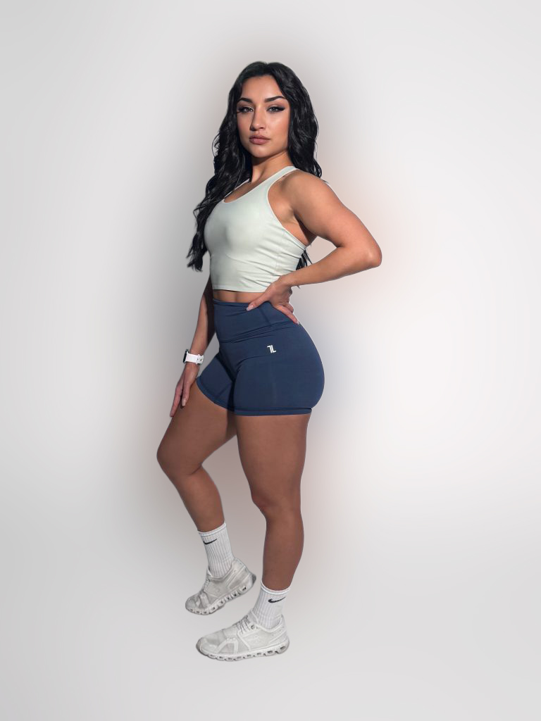 A woman with long dark hair confidently poses in a FlowEase BraTop, blue shorts, white socks, and white sneakers against a plain white background.