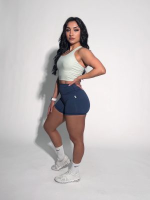 A woman with long dark hair confidently poses in a FlowEase BraTop, blue shorts, white socks, and white sneakers against a plain white background.