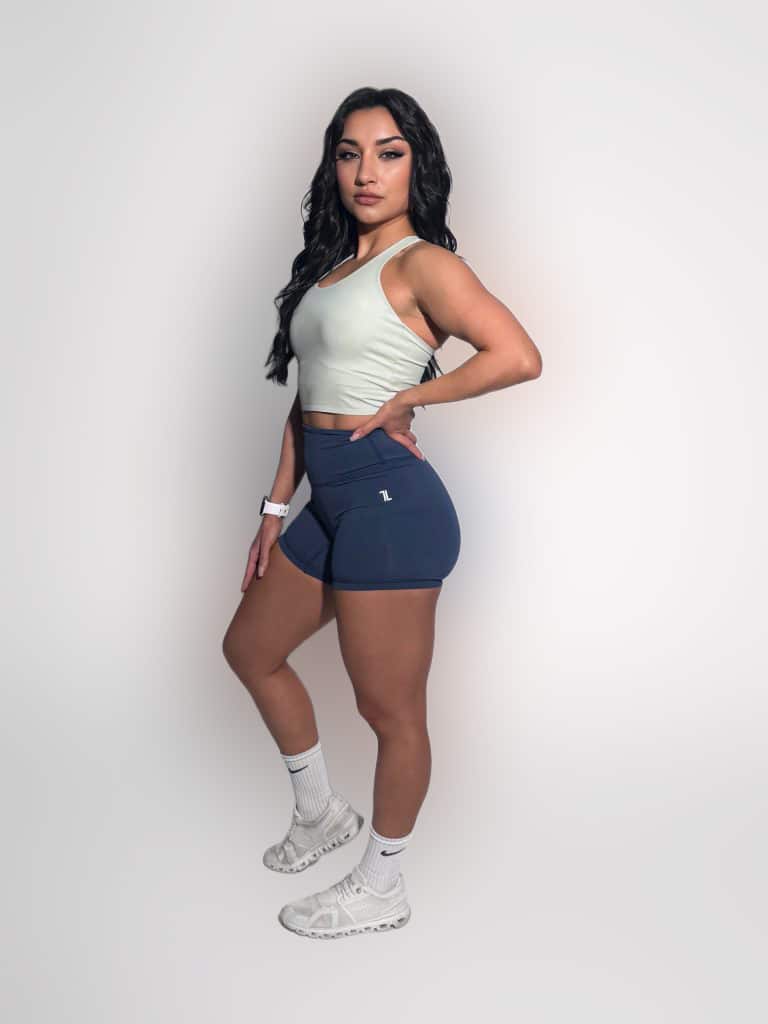 A woman with long dark hair confidently poses in a FlowEase BraTop, blue shorts, white socks, and white sneakers against a plain white background.