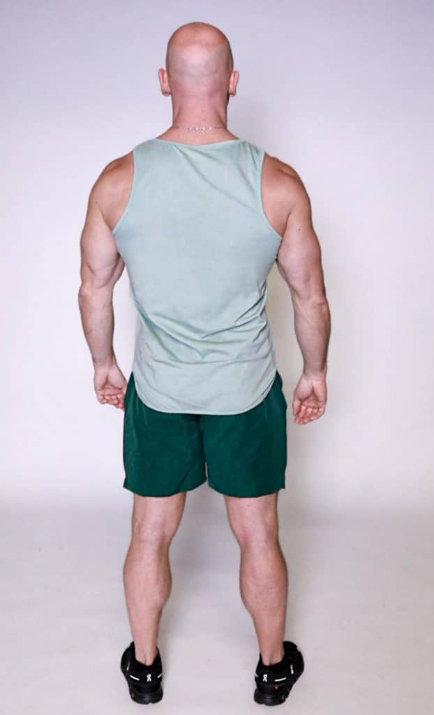 A muscular, bald man faces away from the camera, wearing the Genesis Tank in light green with dark green shorts and black athletic shoes, set against a plain light background.