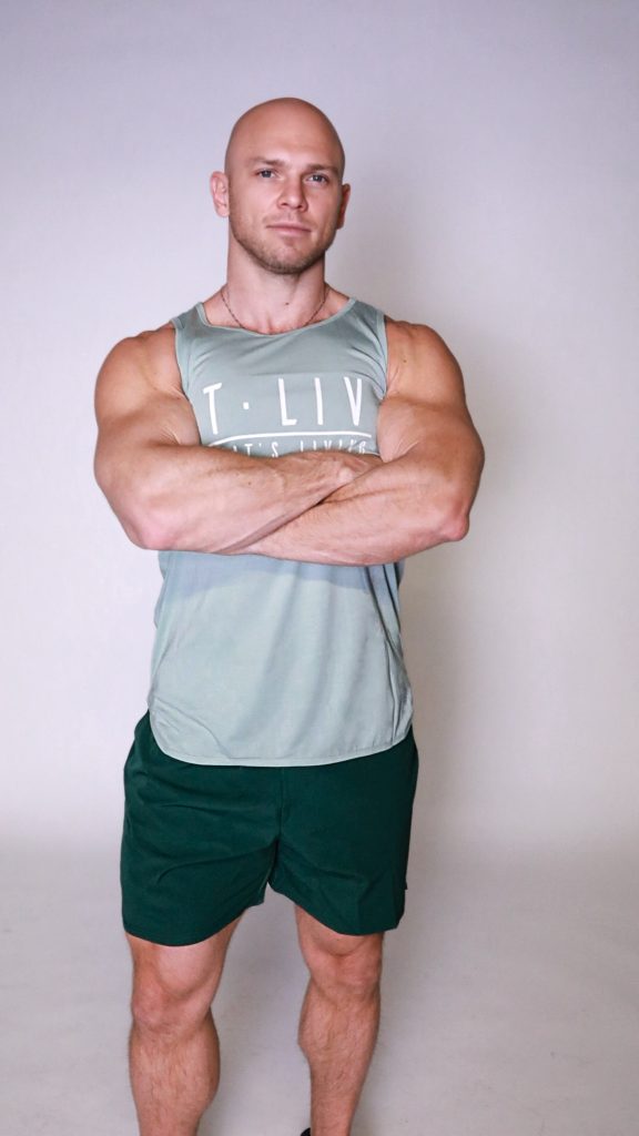 A muscular, bald man stands confidently against a light background, wearing a grey Genesis Tank and dark shorts, with arms crossed.