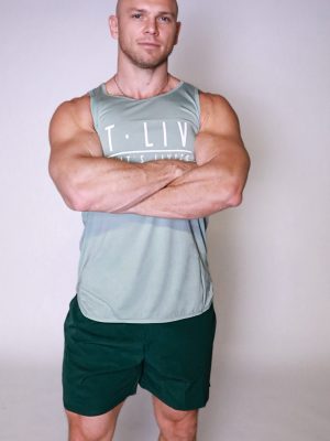 A muscular, bald man stands confidently against a light background, wearing a grey Genesis Tank and dark shorts, with arms crossed.