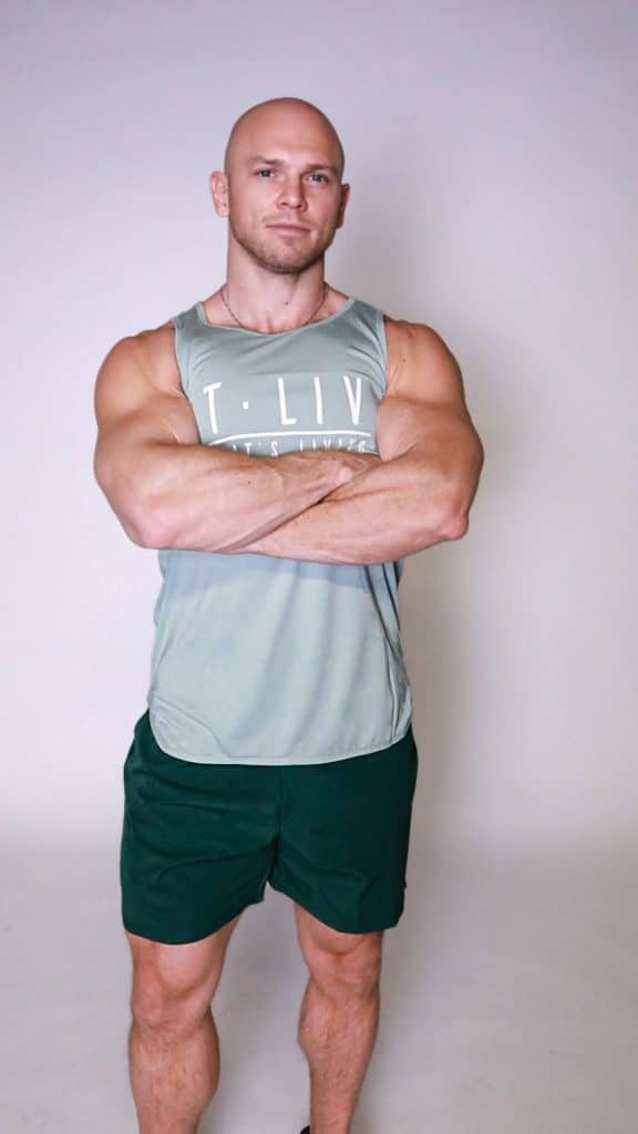 A muscular, bald man stands confidently against a light background, wearing a grey Genesis Tank and dark shorts, with arms crossed.