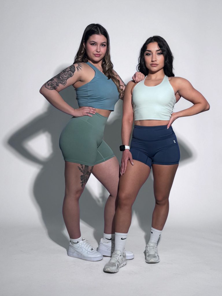 Two women with athletic builds pose confidently in a studio wearing the FlowEase BraTop—one in blue with green shorts, the other in light with dark shorts. Both complete their look with white socks and sneakers.