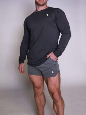 A man with a shaved head is smiling, standing with one hand in his pocket. He is wearing a long-sleeve dark shirt, gray athletic shorts, and black sneakers, posing against a plain light background.