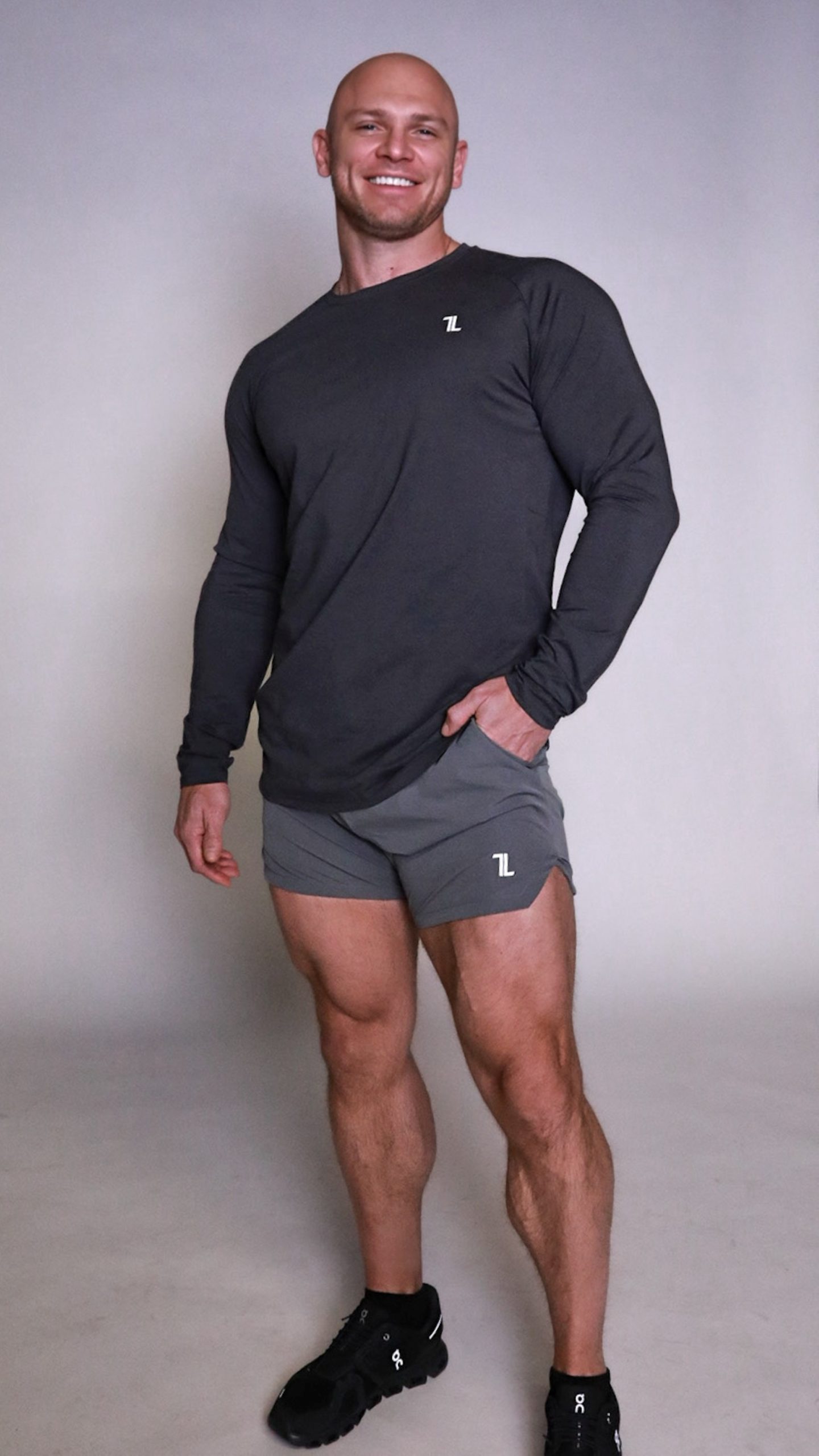 A man with a shaved head is smiling, standing with one hand in his pocket. He is wearing a long-sleeve dark shirt, gray athletic shorts, and black sneakers, posing against a plain light background.