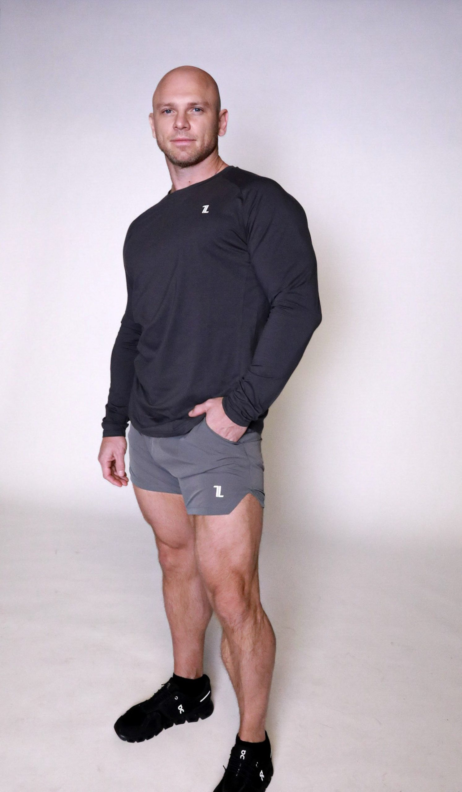 A bald man wearing a long-sleeve dark athletic shirt, gray shorts, and black sneakers stands facing forward against a plain light background.