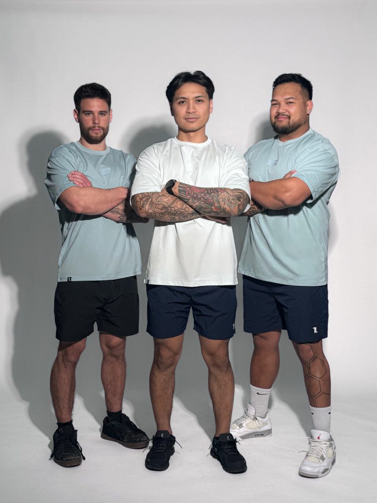 Three men pose confidently in Equity Tees—men’s performance shirts with a tapered fit and scoop bottom—paired with dark shorts. The center man folds his arms while the others stand with arms crossed, all against a plain white background.