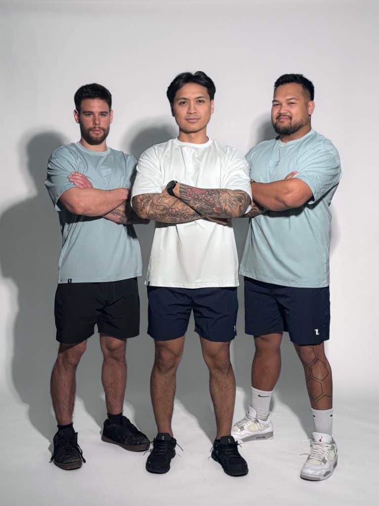 Three men pose confidently in Equity Tees—men’s performance shirts with a tapered fit and scoop bottom—paired with dark shorts. The center man folds his arms while the others stand with arms crossed, all against a plain white background.