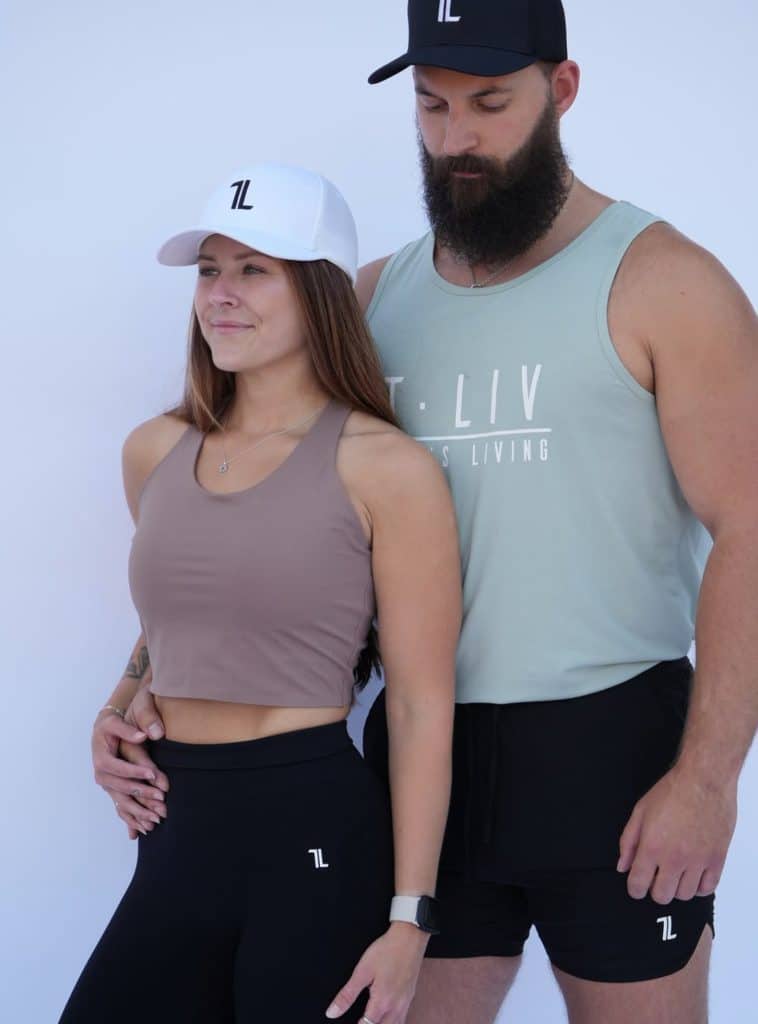 A woman and man wearing Genesis Tank athletic tops and matching hats stand side by side against a white background, both with neutral expressions. The man, who has a beard, rests his hand on the woman's waist.