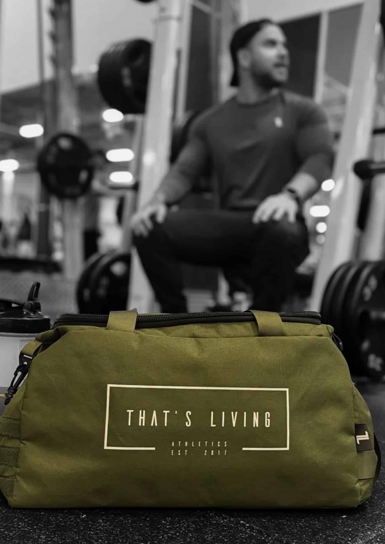 A TRIARC Duffle Bag in green with “THAT’S LIVING ATHLETICS EST 2017” printed on it sits in focus on the gym floor, while a man in workout attire is seen near weightlifting equipment in the blurred background.