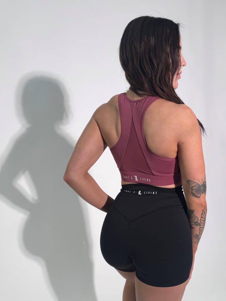 A woman with long dark hair stands facing away in a mauve sports bra and black shorts against a light background, with her left hand on her hip and her shadow cast on the wall.
