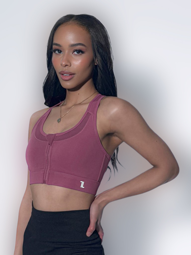A woman with long dark hair wears a mauve sports bra and black leggings, standing with one hand on her hip against a plain light background.