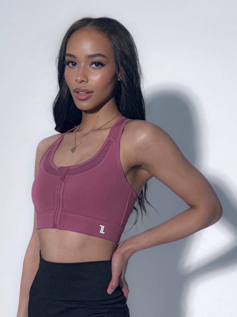 A woman with long dark hair wears a mauve sports bra and black leggings, standing with one hand on her hip against a plain light background.