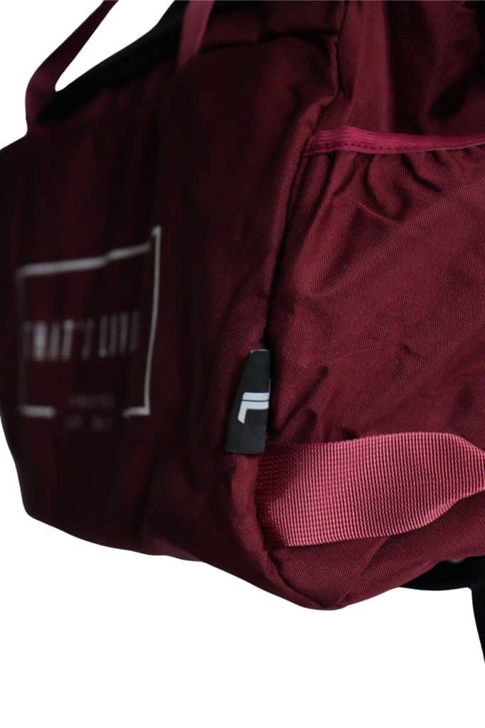 A close-up of a maroon TRIARC Duffle Bag highlighting the side pocket, handle, and part of the brand logo tag on the edge.