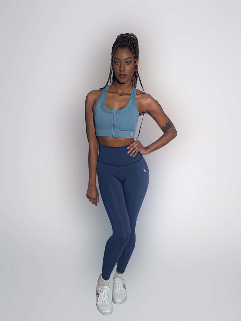 A woman with long braided hair stands confidently against a plain background, wearing a blue zip-up sports bra, matching high-waisted leggings, and white sneakers. Her left arm rests by her side, and she has a tattoo on her upper arm.