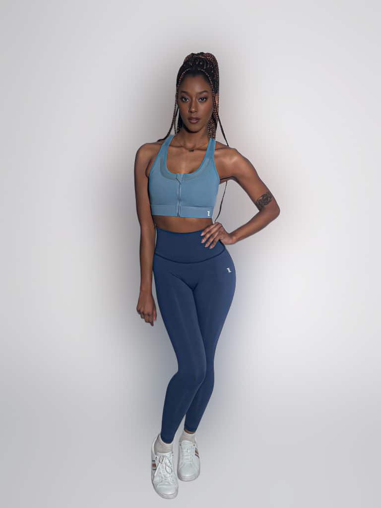 A woman with long braided hair stands confidently against a plain background, wearing a blue zip-up sports bra, matching high-waisted leggings, and white sneakers. Her left arm rests by her side, and she has a tattoo on her upper arm.
