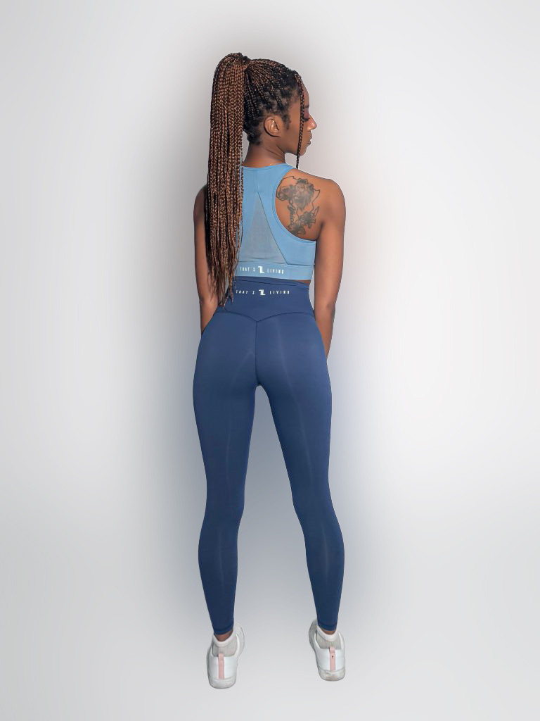 A woman with long braided hair, wearing a blue sleeveless athletic top and matching leggings, stands facing away in a well-lit studio. A tattoo is visible on her left shoulder, and she is wearing white sneakers.
