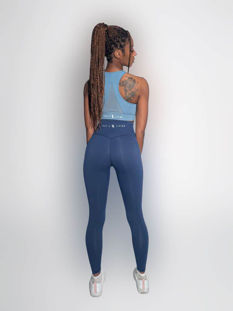 A woman with long braided hair, wearing a blue sleeveless athletic top and matching leggings, stands facing away in a well-lit studio. A tattoo is visible on her left shoulder, and she is wearing white sneakers.