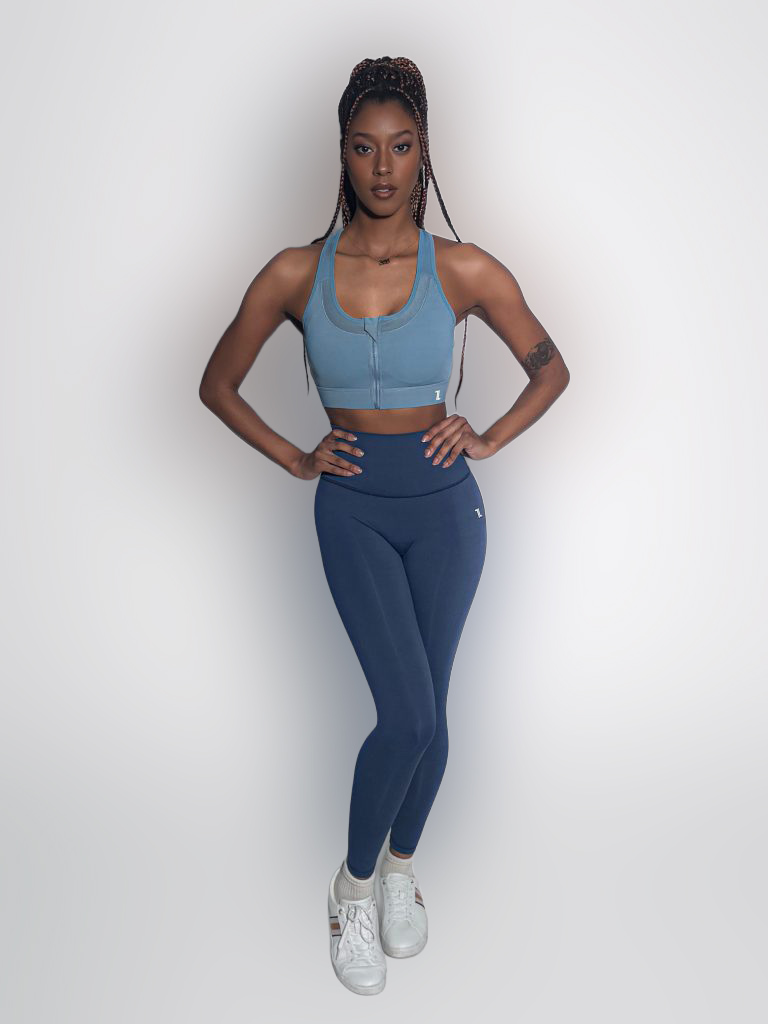 A woman stands confidently against a plain white background, wearing a blue sports bra, matching blue leggings, and white sneakers. She has long braided hair and rests her hands on her hips.