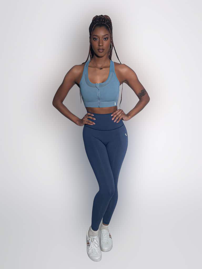 A woman stands confidently against a plain white background, wearing a blue sports bra, matching blue leggings, and white sneakers. She has long braided hair and rests her hands on her hips.