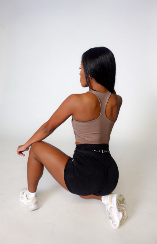 A woman with long black hair, dressed in a brown tank top and Contour Shorts, kneels on one knee facing away from the camera against a plain white background.