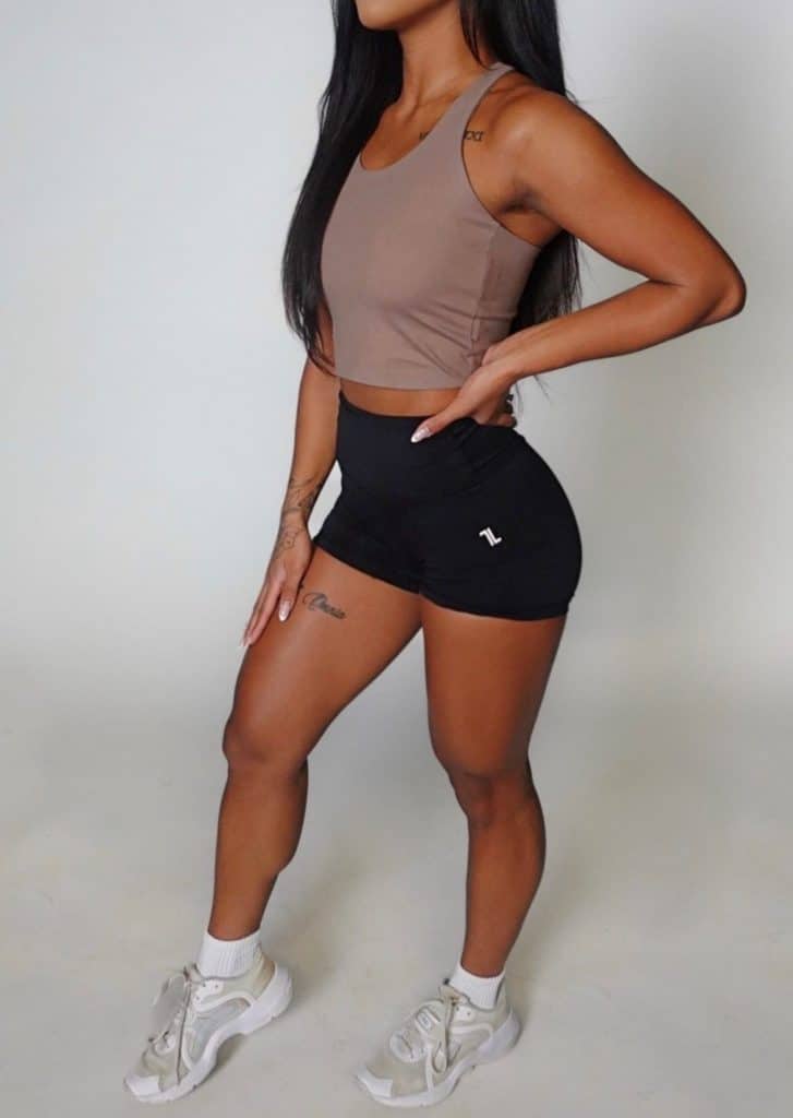 A woman in a taupe crop tank top, black high-waisted shorts, white socks, and white sneakers stands in a relaxed pose against a plain white background. Her long hair is down and her left hand rests on her hip.