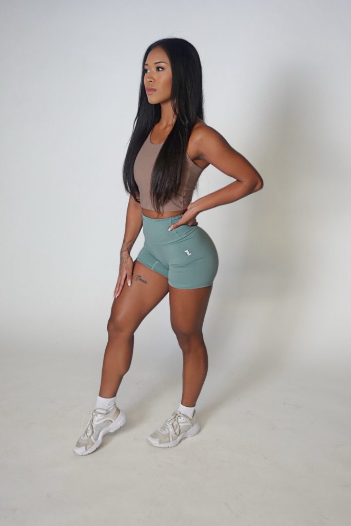 A woman with long black hair stands confidently in a studio, wearing a brown crop top, teal athletic shorts, white socks, and white sneakers against a plain white background.