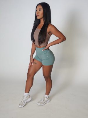 A woman with long black hair stands confidently in a studio, wearing a brown crop top, teal athletic shorts, white socks, and white sneakers against a plain white background.