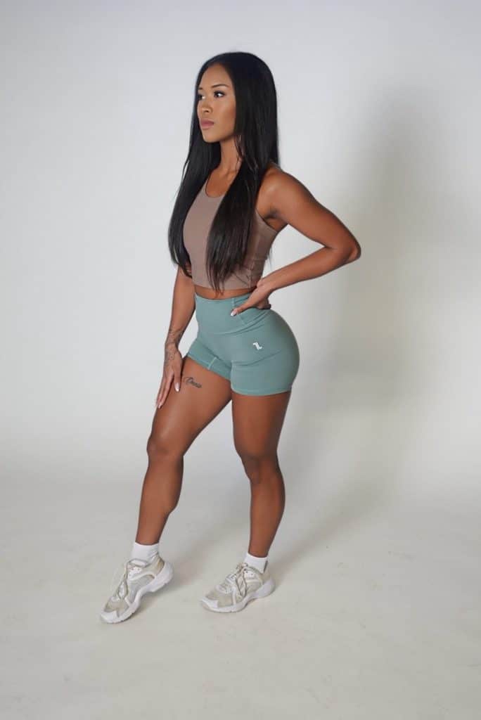 A woman with long black hair stands confidently in a studio, wearing a brown crop top, teal athletic shorts, white socks, and white sneakers against a plain white background.