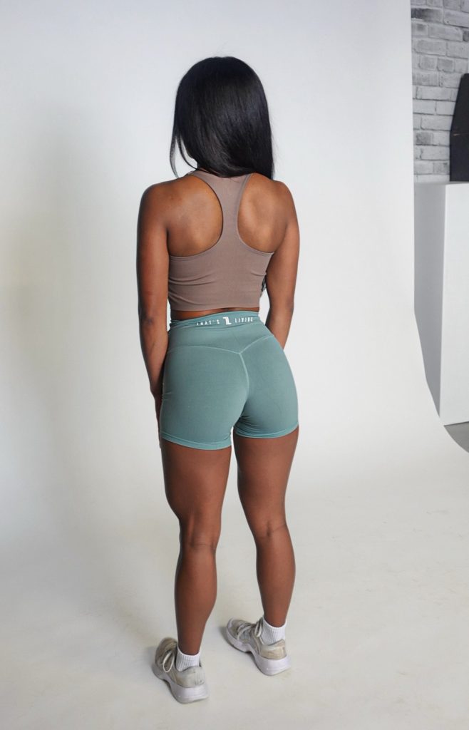 A woman with long dark hair stands facing away, wearing a brown sports bra, teal athletic shorts, and white sneakers in a photo studio with a white backdrop.