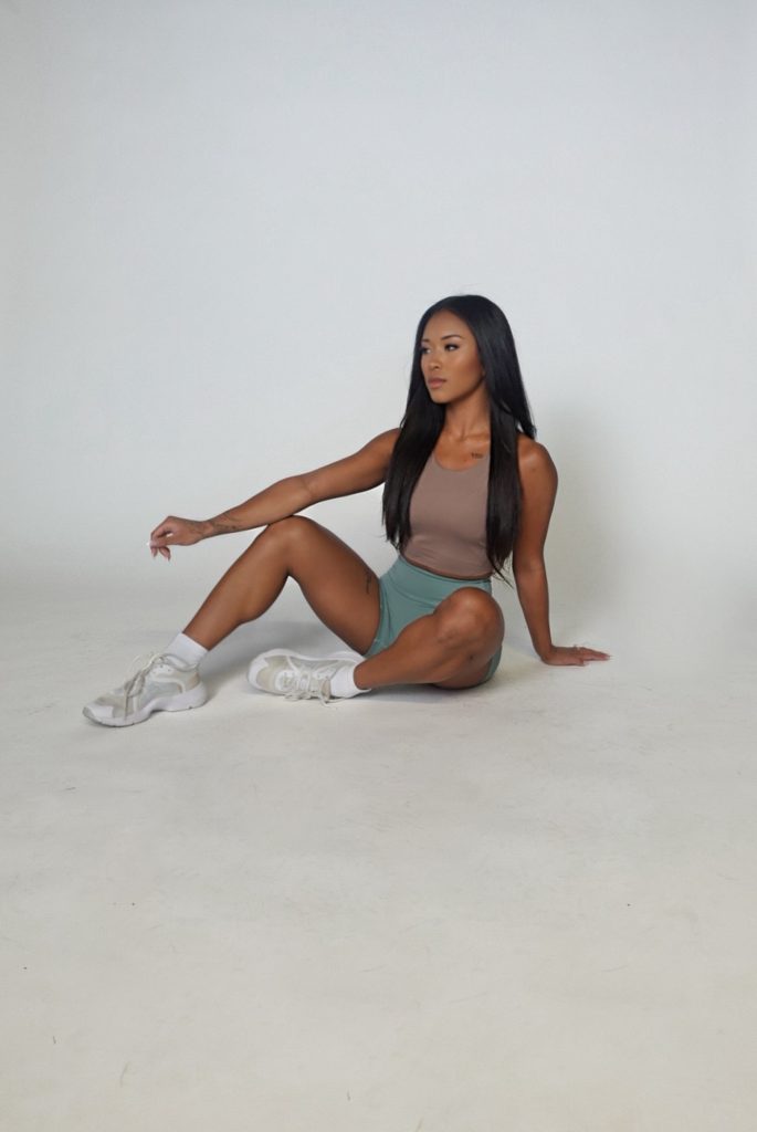 A woman with long dark hair sits on the floor against a plain white background, wearing a beige tank top, teal shorts, and white sneakers. One arm rests on her knee while the other is placed on the ground for support.