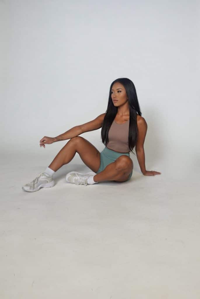 A woman with long dark hair sits on the floor against a plain white background, wearing a beige tank top, teal shorts, and white sneakers. One arm rests on her knee while the other is placed on the ground for support.