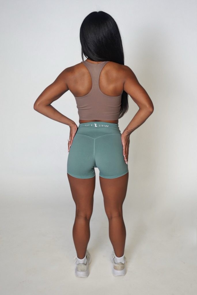 A woman with long dark hair stands facing away, wearing a brown sports bra, green athletic shorts, and white sneakers, against a plain light background.