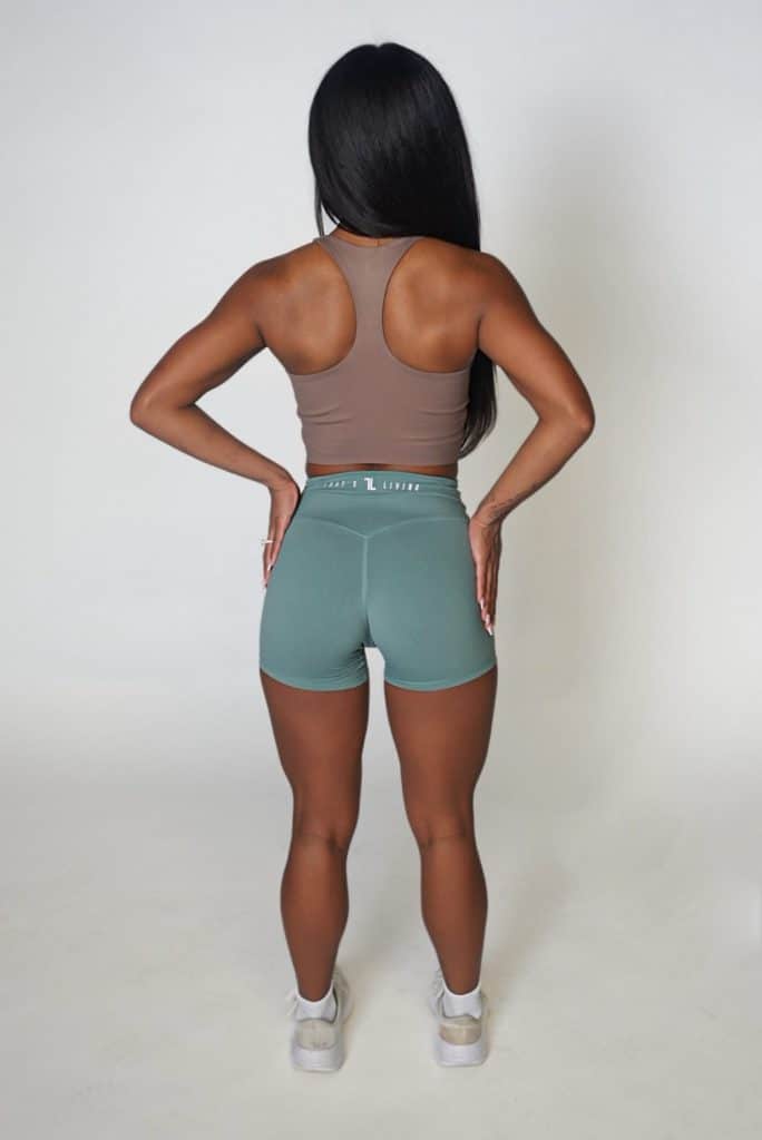 A woman with long dark hair stands facing away, wearing a brown sports bra, green athletic shorts, and white sneakers, against a plain light background.
