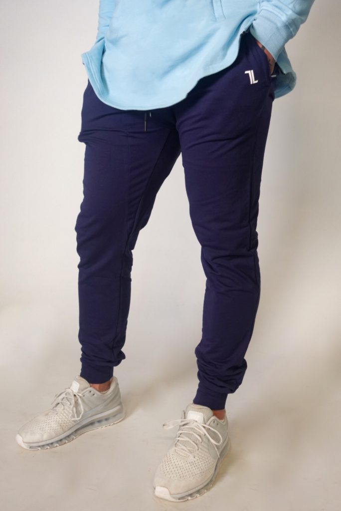 A person wearing a light blue hoodie, dark blue jogger pants, and light gray athletic shoes stands against a plain background. The person's hands are in their hoodie pockets.