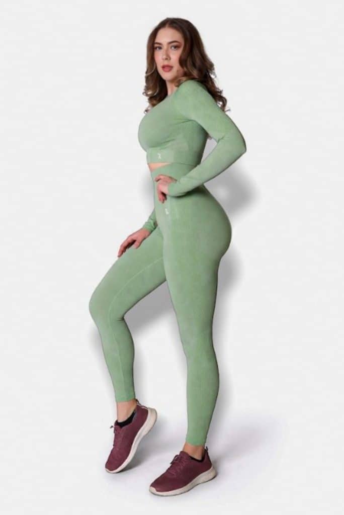 A woman wearing a light green, long-sleeve crop top and matching leggings poses standing sideways. She is wearing maroon athletic shoes and is set against a plain white background.