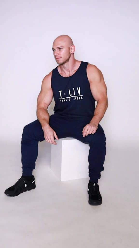 A muscular man in a navy tank top and pants sits on a white cube against a plain white background. He is looking to the side with one hand resting on his knee.