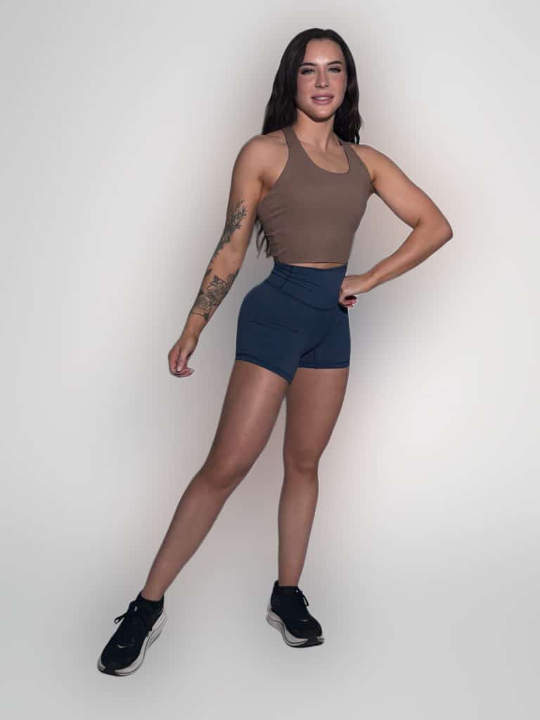 A woman with long dark hair poses confidently against a plain light background, wearing a brown sleeveless crop top, navy blue athletic shorts, and black sneakers. She has a tattoo on her left arm.