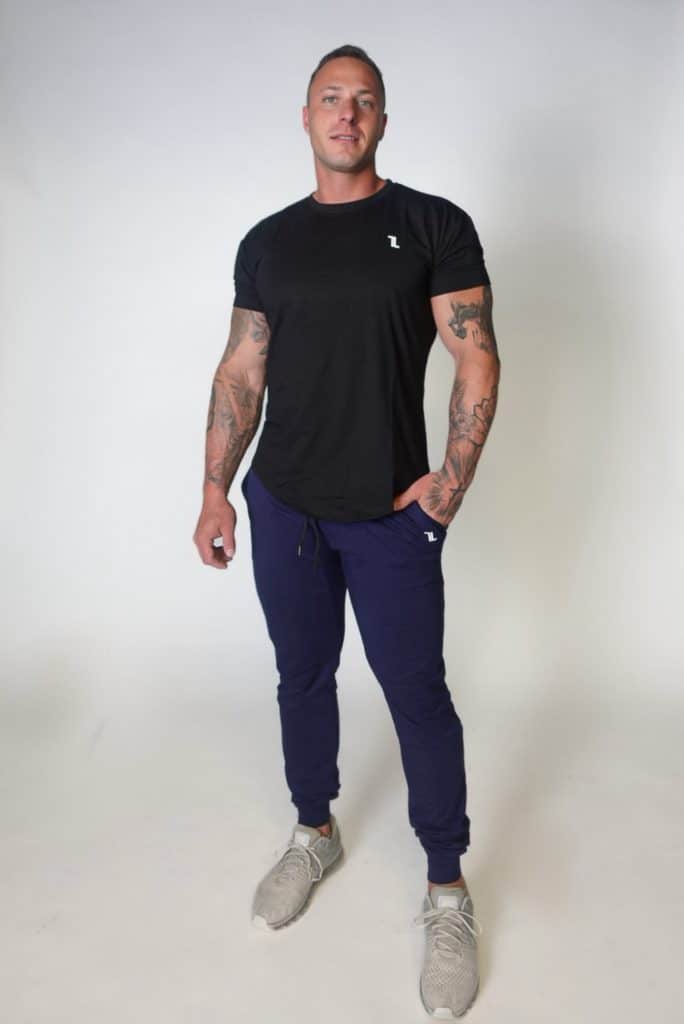 A man with tattoos stands against a plain white background, wearing a black T-shirt, navy joggers, and gray sneakers. One hand is in his pocket and he looks directly at the camera.