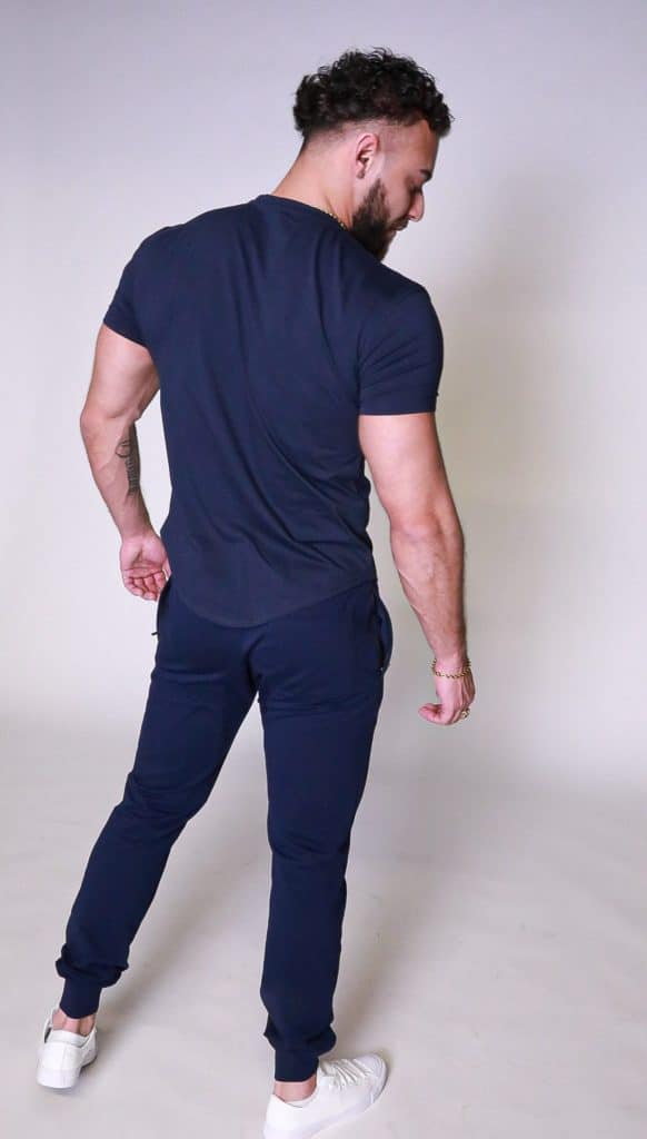 A man with short dark hair and a beard stands facing away from the camera, wearing a navy blue t-shirt, matching joggers, and white sneakers against a light background.