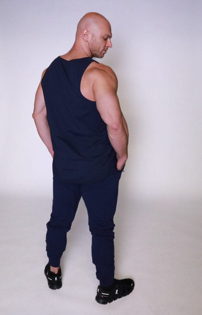 A muscular, bald man wearing a navy blue tank top, matching navy pants, and black sneakers stands with his back mostly to the camera, head slightly turned, in a plain, well-lit studio setting.
