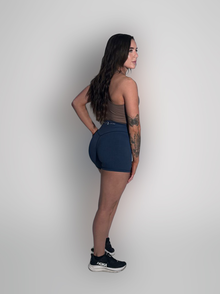 A woman with long brown hair, wearing a brown sports bra, navy blue shorts, and black Hoka sneakers, stands in profile against a plain light background, showing tattoos on her left arm.