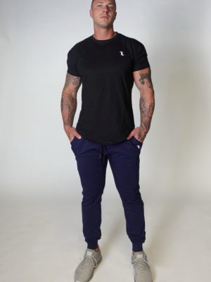 A man with tattoos stands against a plain light background, wearing a black t-shirt, navy blue jogger pants, and light gray sneakers, with his hands in his pockets and a neutral expression.