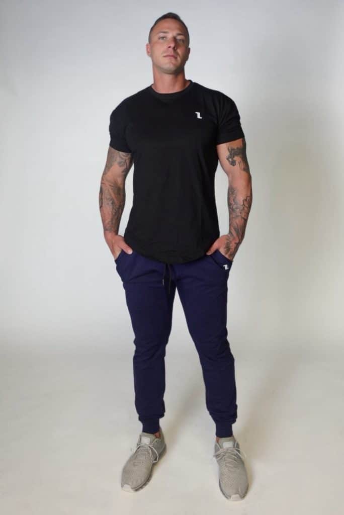 A man with tattoos stands against a plain light background, wearing a black t-shirt, navy blue jogger pants, and light gray sneakers, with his hands in his pockets and a neutral expression.