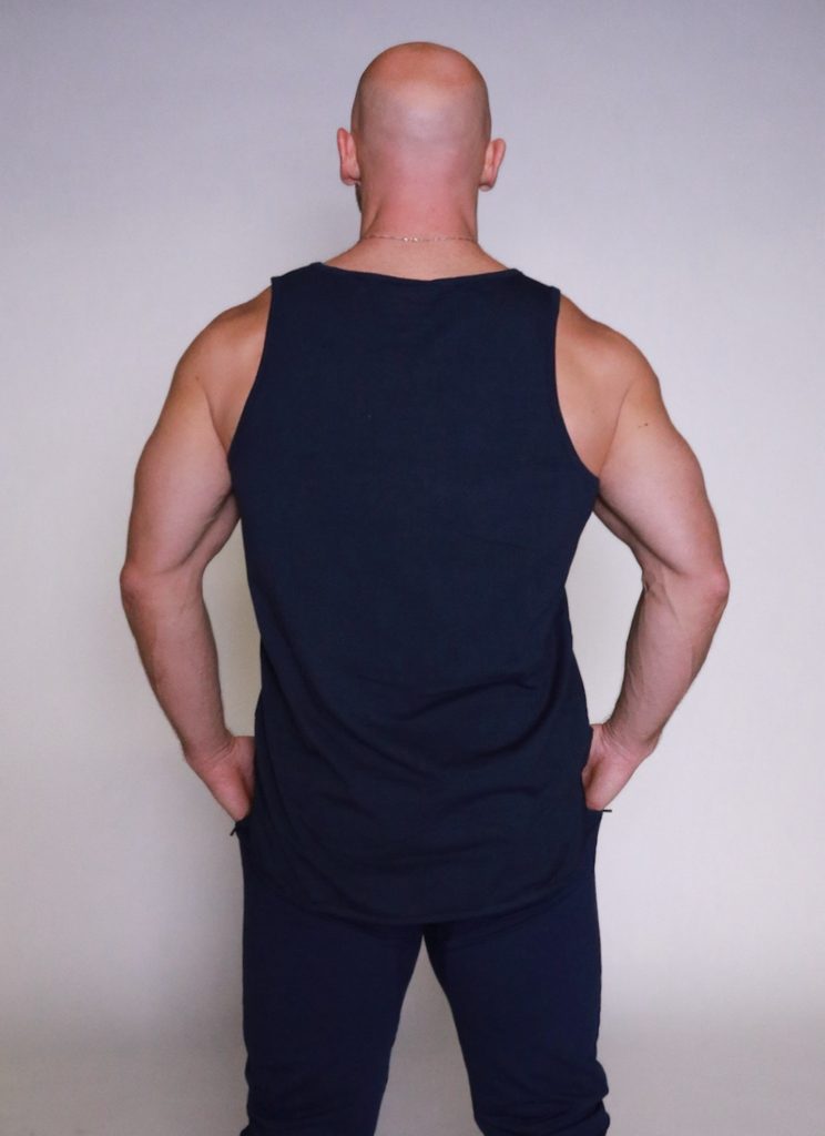 A bald man wearing a black tank top and black pants stands facing away from the camera with his hands resting on his hips against a plain light background.