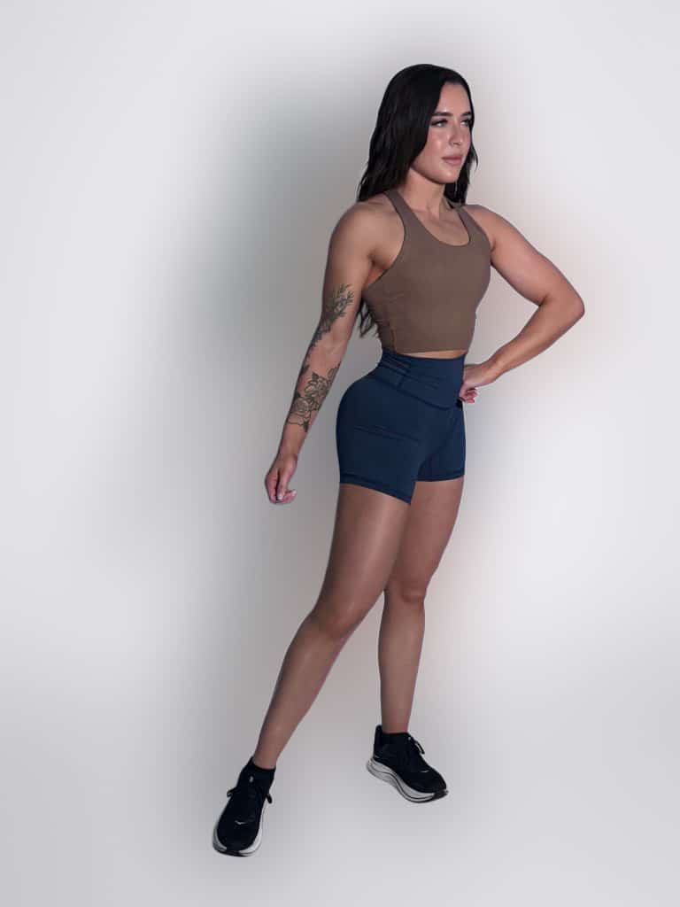 A woman in athletic wear stands confidently against a plain light background. She wears a brown tank top, navy shorts, and black sneakers, with her right hand on her hip and a tattoo visible on her left arm.