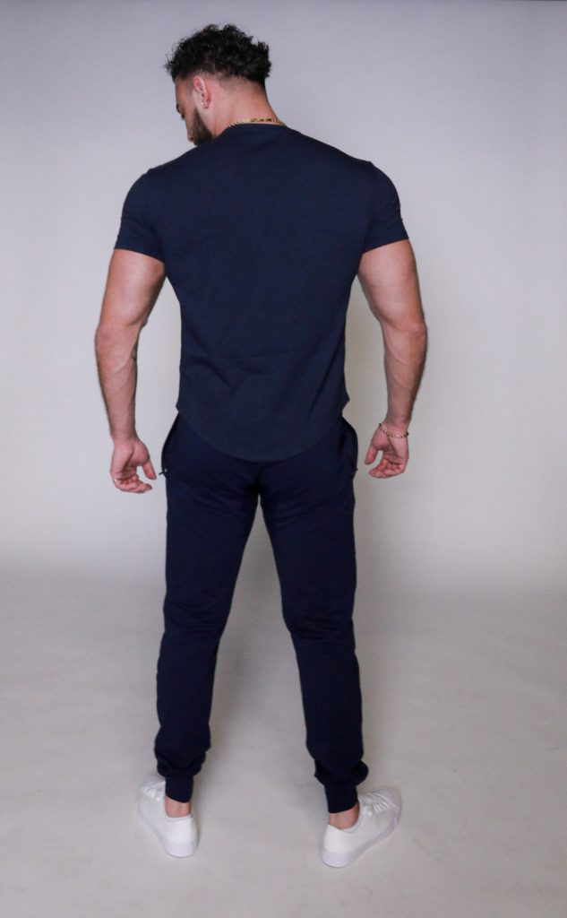 A man with short dark hair stands facing away from the camera. He is wearing a fitted dark blue t-shirt, dark blue pants, and white sneakers, against a plain light background.
